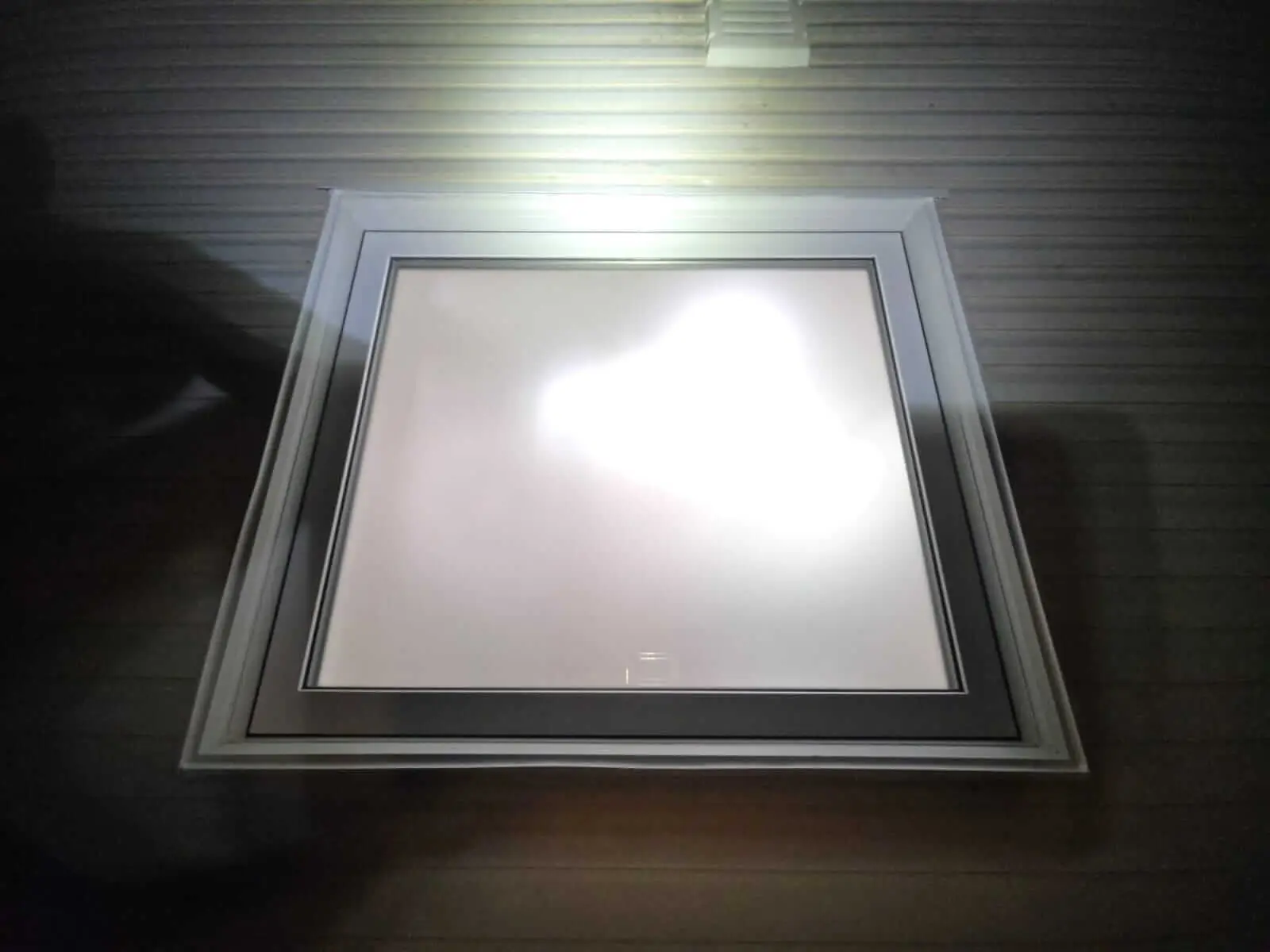 NTI Windows & Doors A square window with a white frame is illuminated by a light source, creating a bright glare in the center. The wall has horizontal lines, complementing the modern style of GTA Doors and windows. Window Replacement Hamilton & GTA