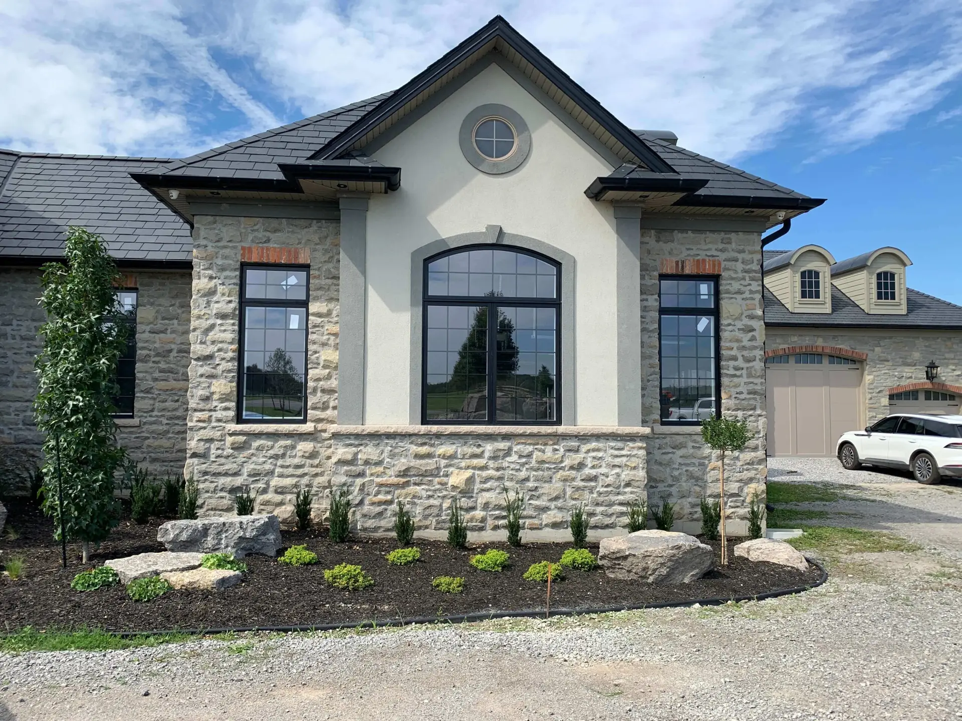 NTI Windows & Doors A stone and stucco house with large black-trimmed GTA Windows, landscaped front yard, and a white car parked on a gravel driveway. Window Replacement Hamilton & GTA