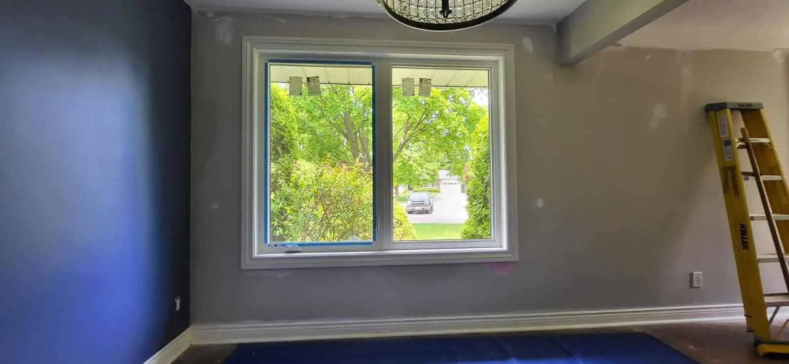 NTI A room with freshly painted blue and gray walls, a large window facing greenery, sliding doors to the left, a ceiling light, and a yellow ladder against the right wall. window and doors