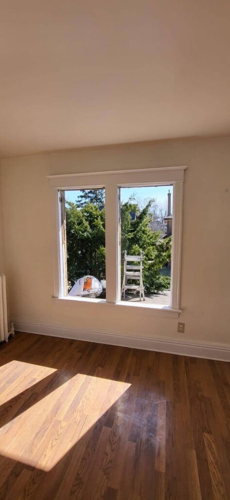 NTI A bright room with hardwood floors and two large windows, one open; outside, a white ladder leans against the house next to trees. window and doors